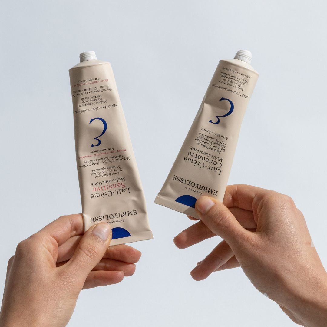 Two tubes of Embryolisse Lait-Creme Concentré, normal size and travel size held by hands against a light gray background
