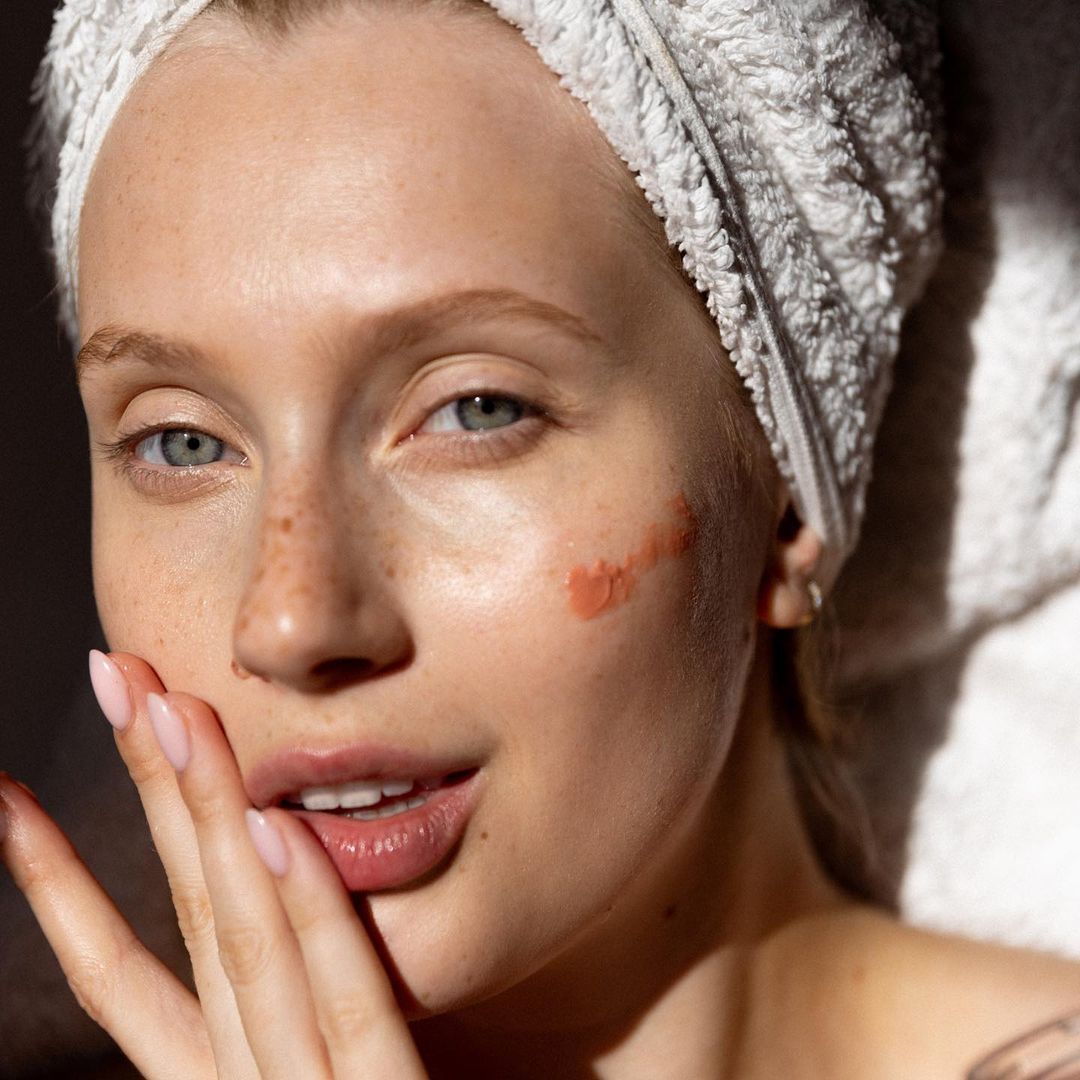 Woman with a towel on her head, showing skin with natural makeup and a pea of Radiant Complexion Cream - Pink Glow skincare product by Embryolisse on her faceñ