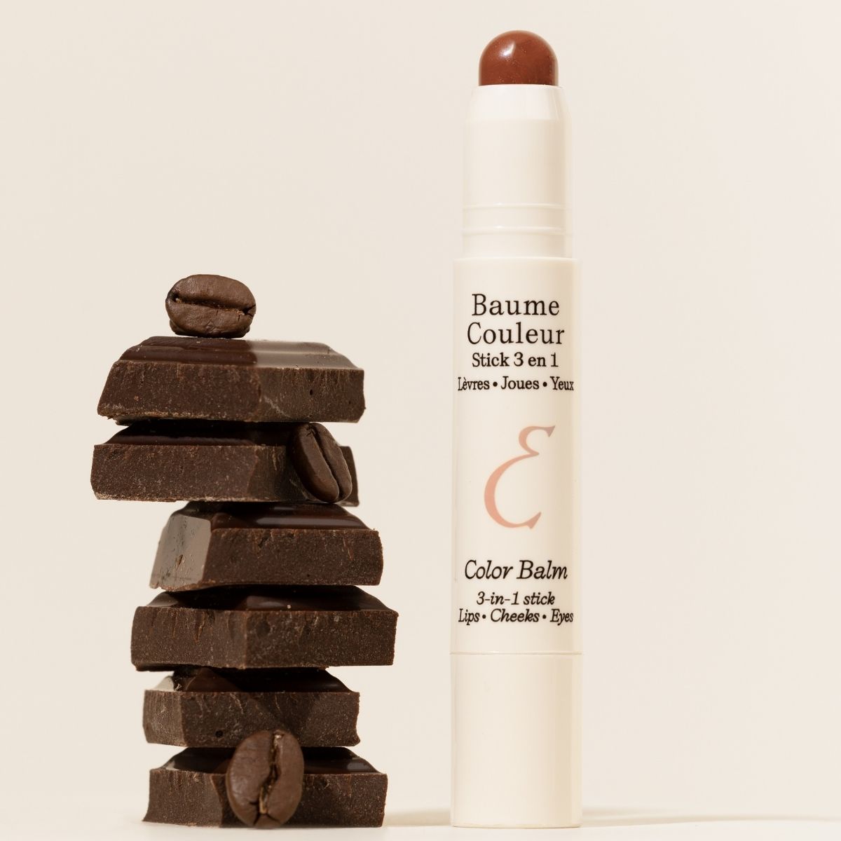 Embryolisse Artist Secret Color Balm 3-in-1 Stick Mocha displayed upright next to stacked chocolate pieces and coffee beans, highlighting the warm brown shade and creamy texture of the multi-use balm designed for lips, cheeks, and eyes
