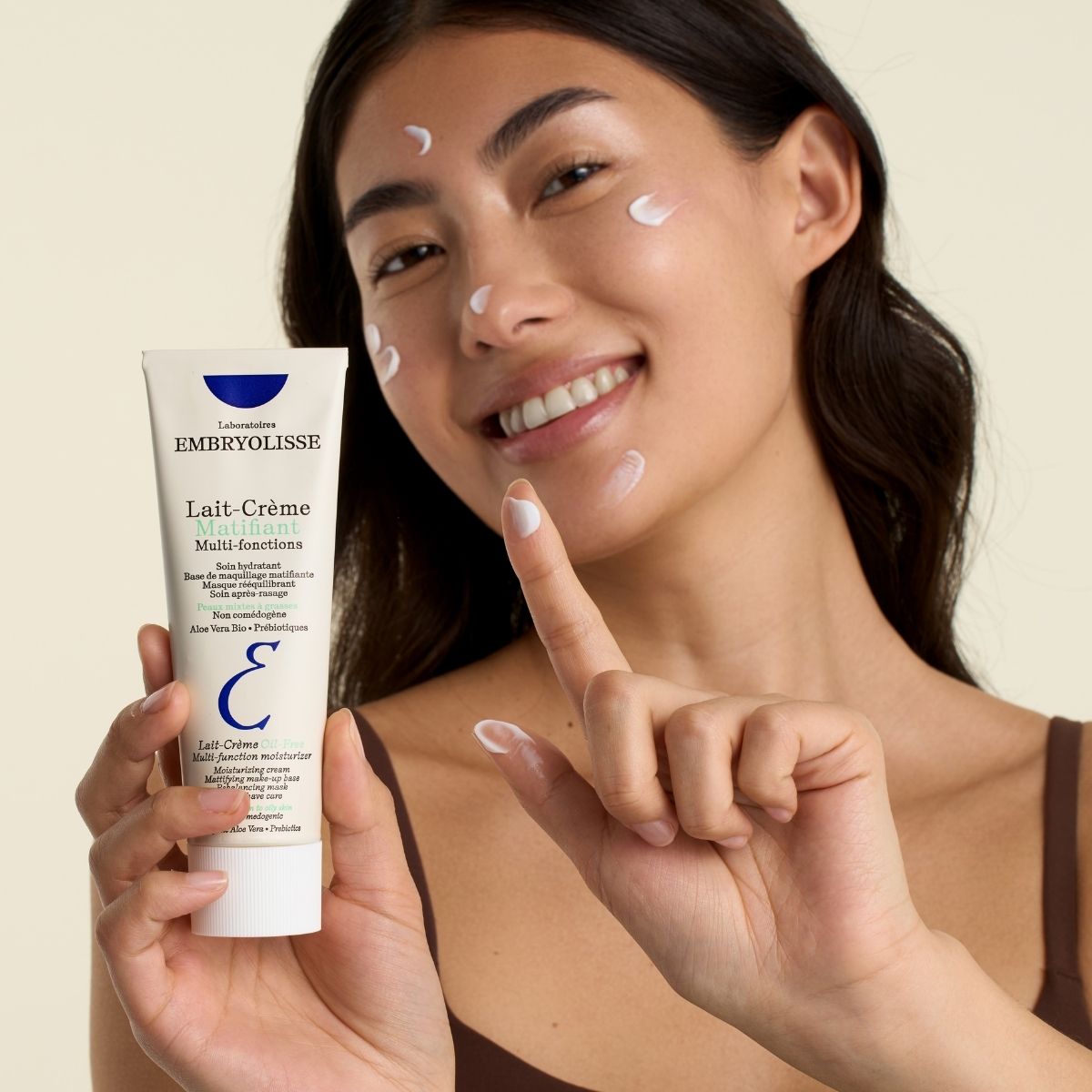 A smiling woman with dots of Lait-Crème Oil-Free on her cheeks and forehead, holding the tube